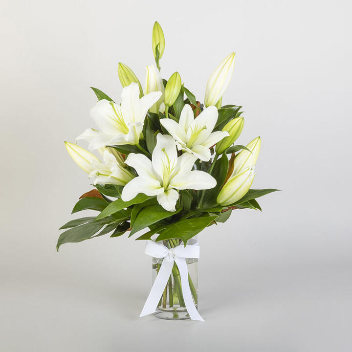  A modern creation of a lily bouquet with greenery arranged in a glass vase. Sympathy lilies' use...